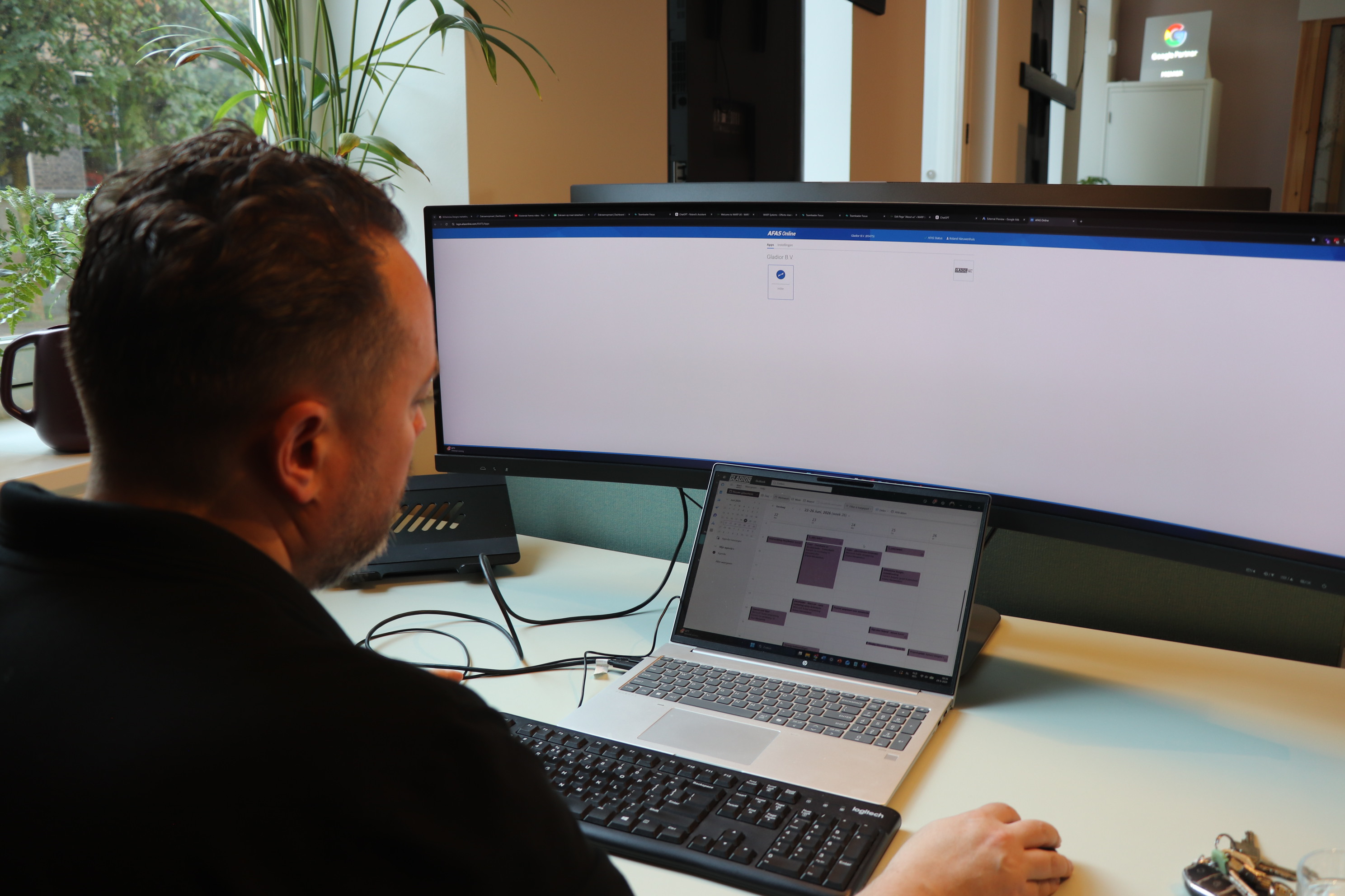 Medewerker werkt achter een breed beeldscherm en laptop aan een AFAS-omgeving en agenda op kantoor.