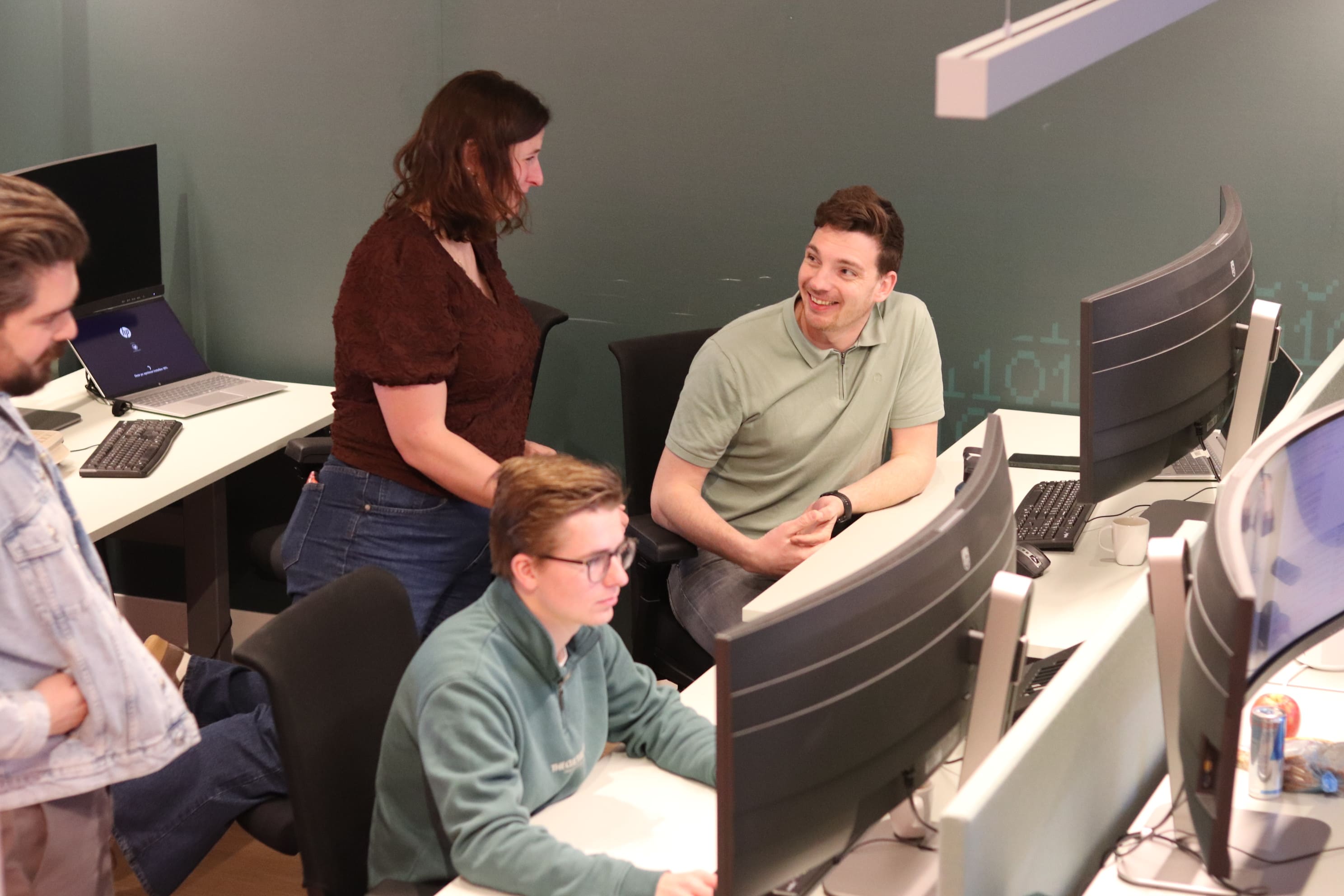 Een groep collega's in overleg bij een werkplek met meerdere computerschermen in een kantooromgeving.