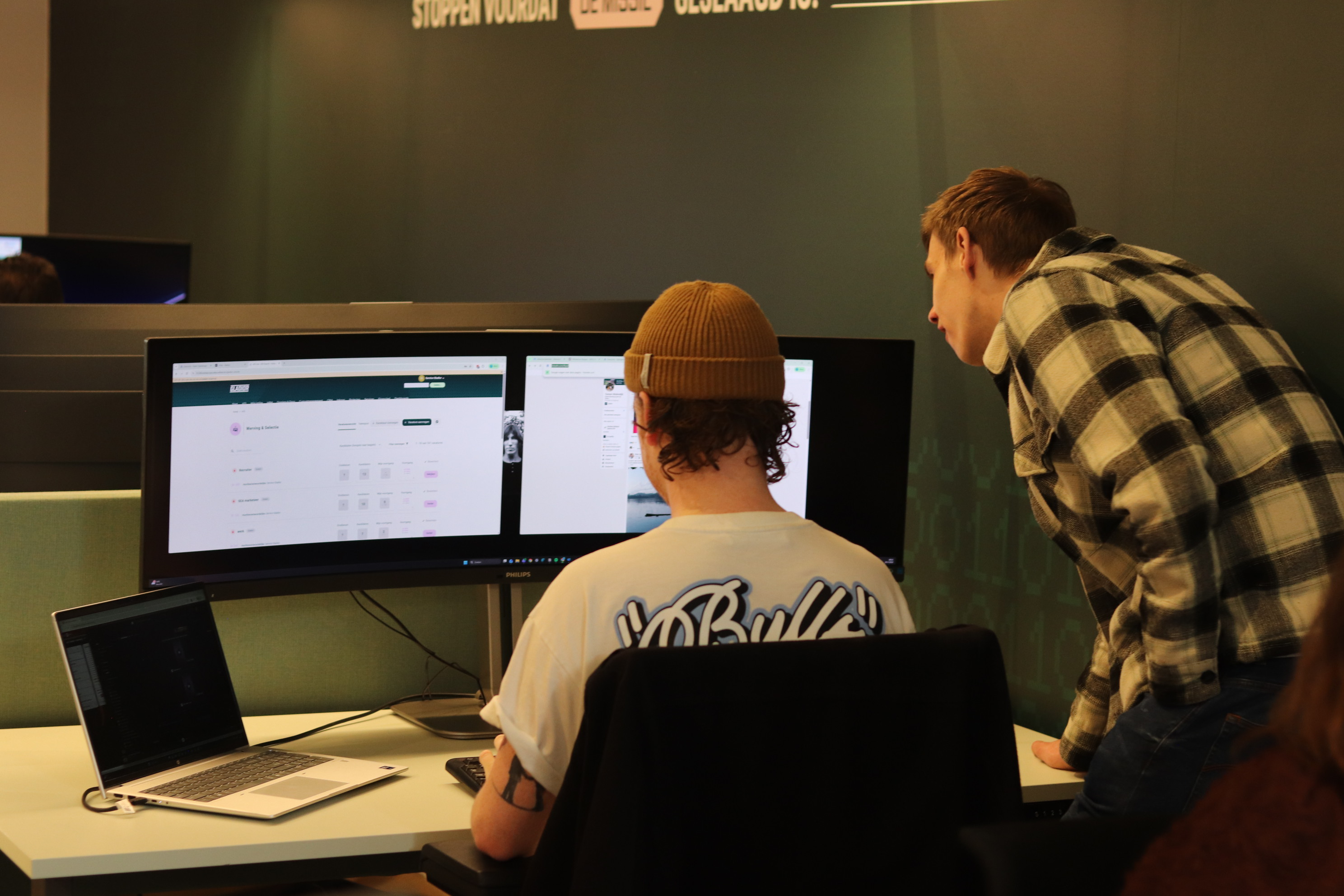 Twee mannen werken samen achter een grote computermonitor op een kantoor. De een zit op een bureaustoel, de ander buigt voorover om mee te kijken naar het scherm. Op het bureau staat ook een laptop.