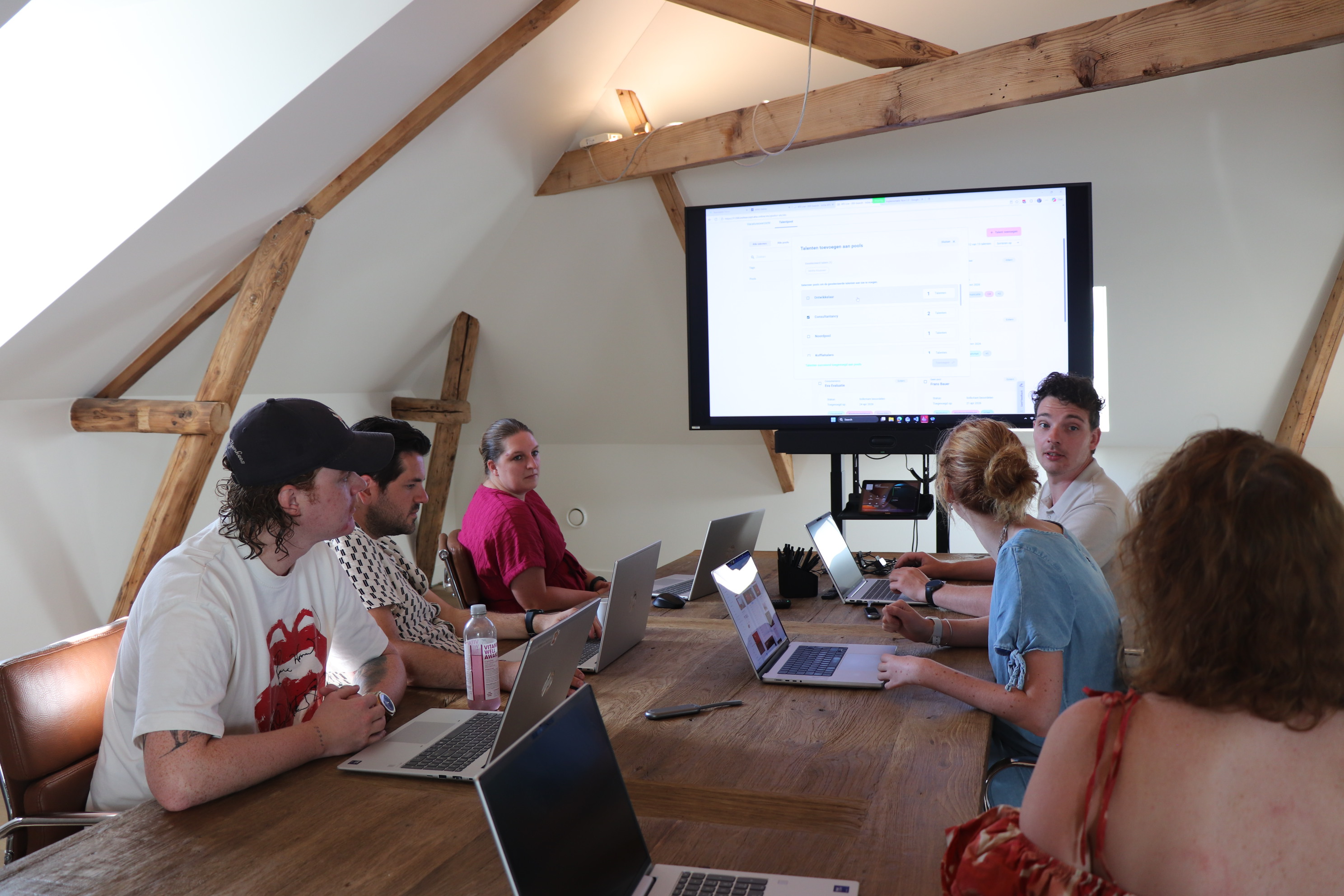 Een groep collega's van NOVI vergadert met laptops aan een houten tafel en kijkt naar een groot presentatiescherm in een lichte kantoorruimte met houten balken.
