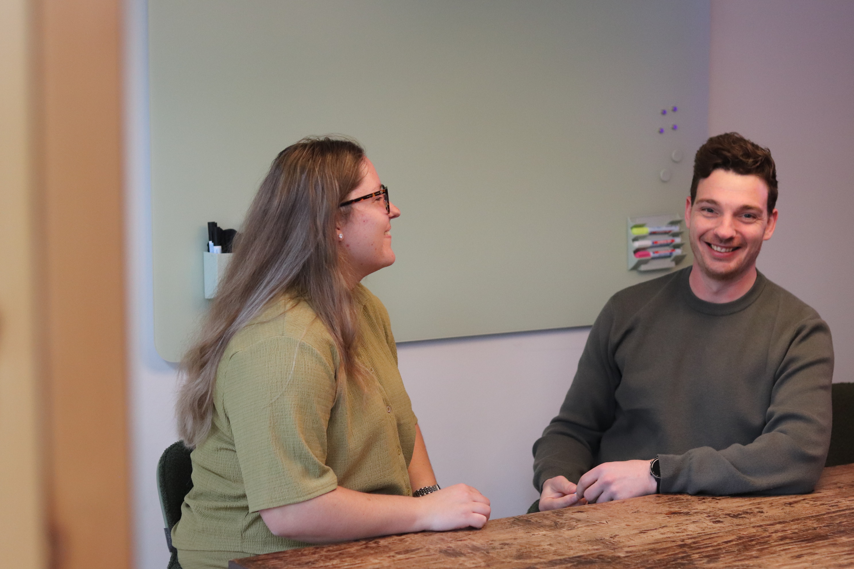 Twee collega’s zitten lachend aan tafel tijdens een informeel overleg op kantoor.