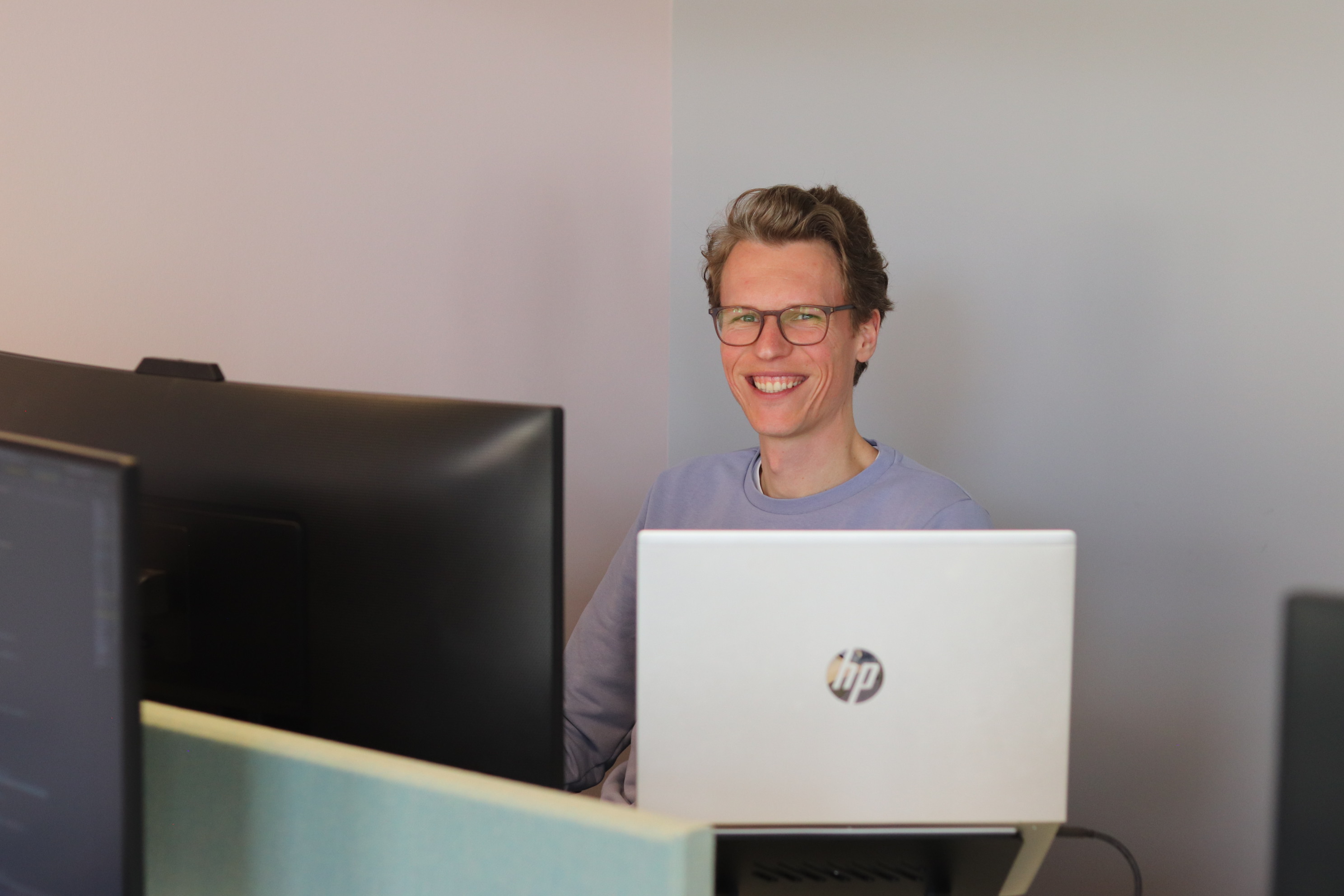 Lachende medewerker werkt achter een laptop en meerdere beeldschermen op kantoor.