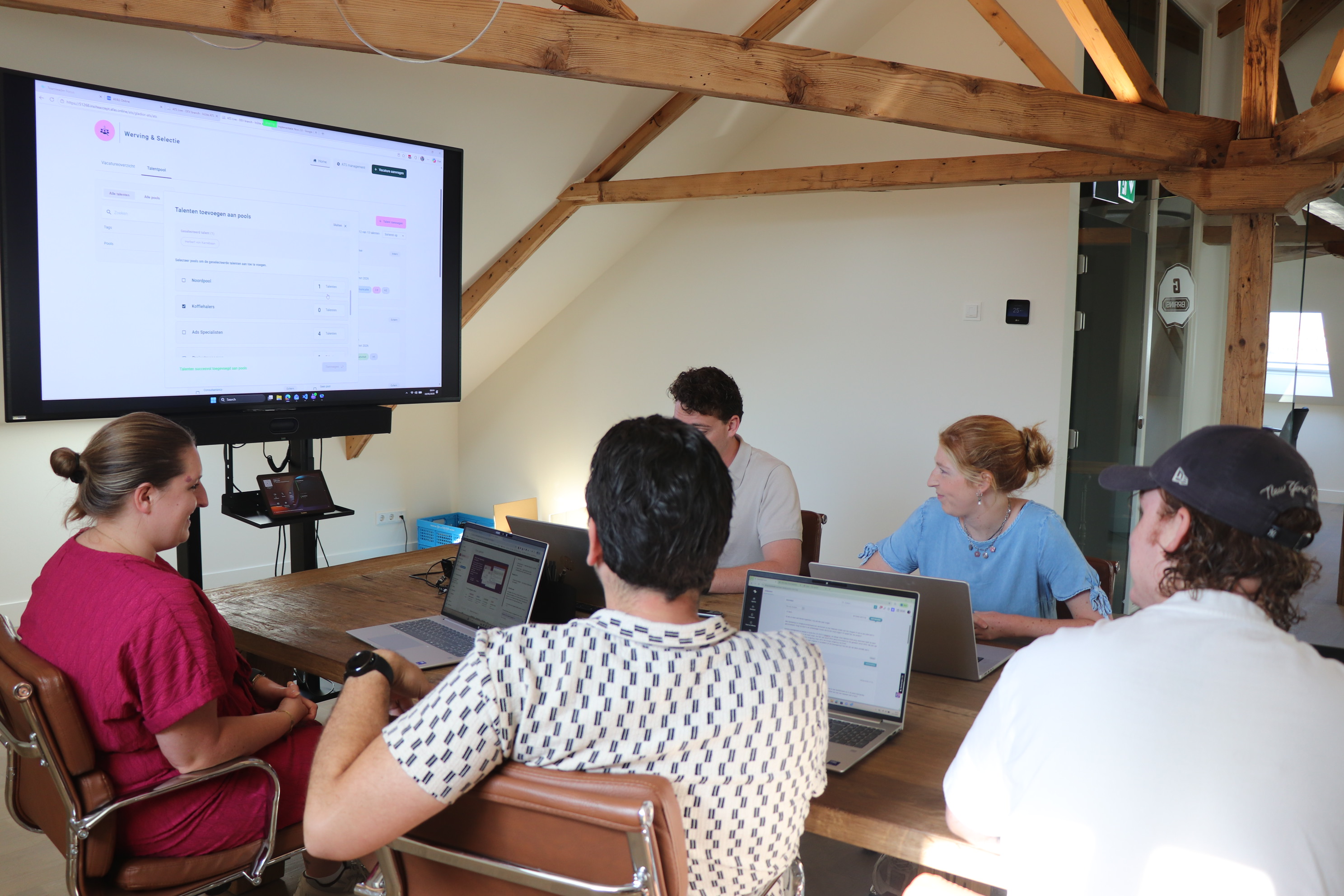 Een groep collega's rond een houten vergadertafel met laptops, kijkend naar een groot presentatiescherm in een kantoorruimte met een houten balkenplafond.