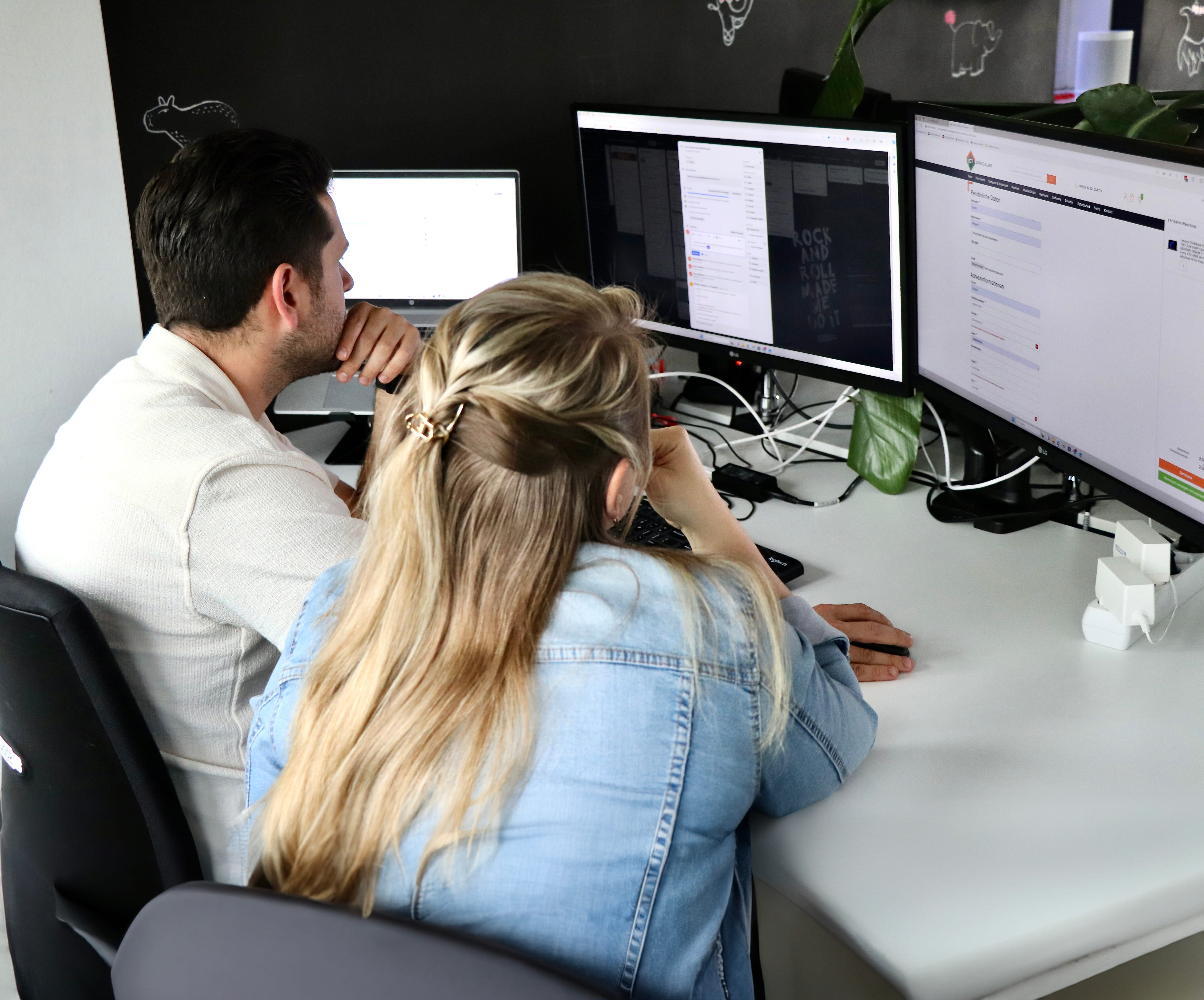 Twee collega's die van achteren gefotografeerd zijn terwijl ze samen geconcentreerd naar meerdere computerschermen kijken op een kantoorwerkplek.